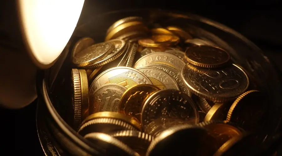 A detailed view of assorted coins in a jar under warm light, depicting financial wealth.