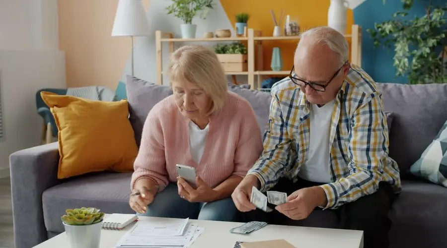 Elderly couple budgeting at home, seated on a cozy sofa, using smartphone and cash.