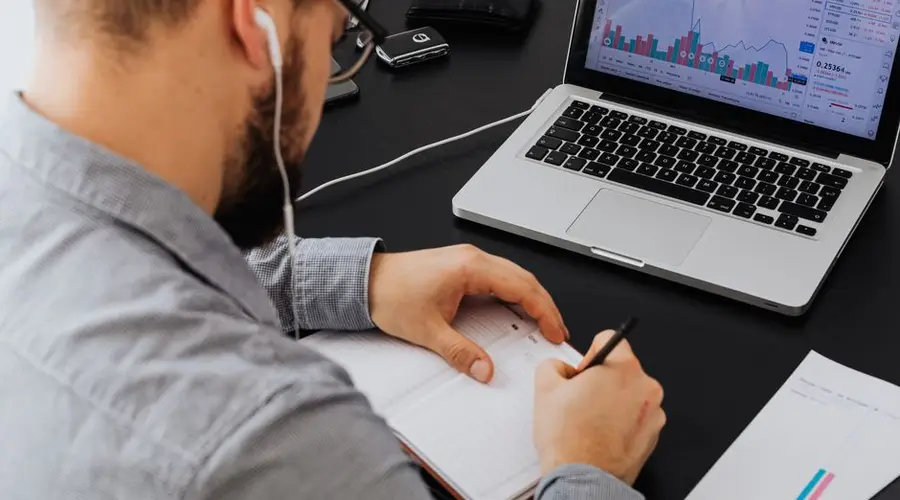 A businessman analyzing stock data on a laptop while taking notes at a desk.