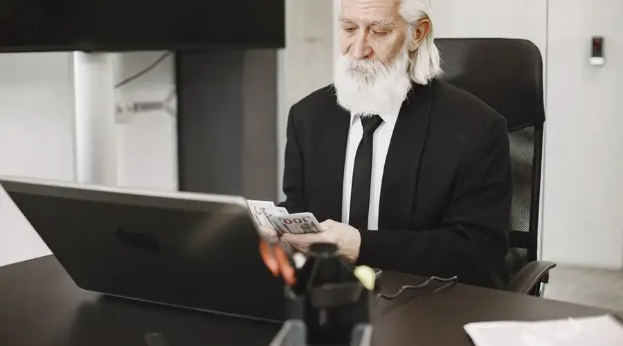 White-haired man in suit counting money at a modern office desk with laptop.