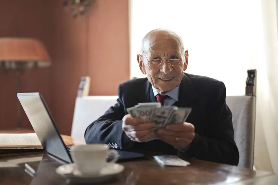 Positive senior businessman in formal suit and eyeglasses holding money in hands while sitting at table near laptop and looking at camera