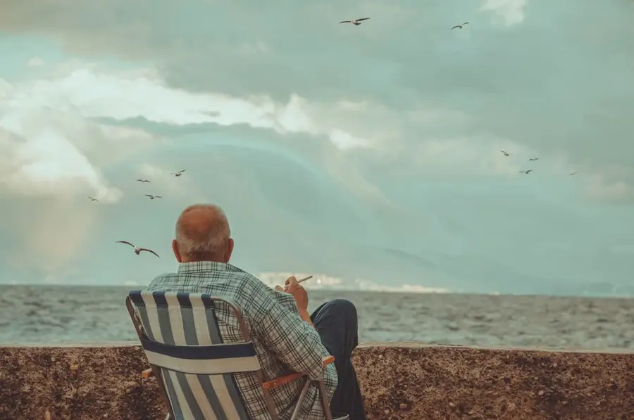 An elderly man sits by the ocean, enjoying a peaceful moment under cloudy skies.