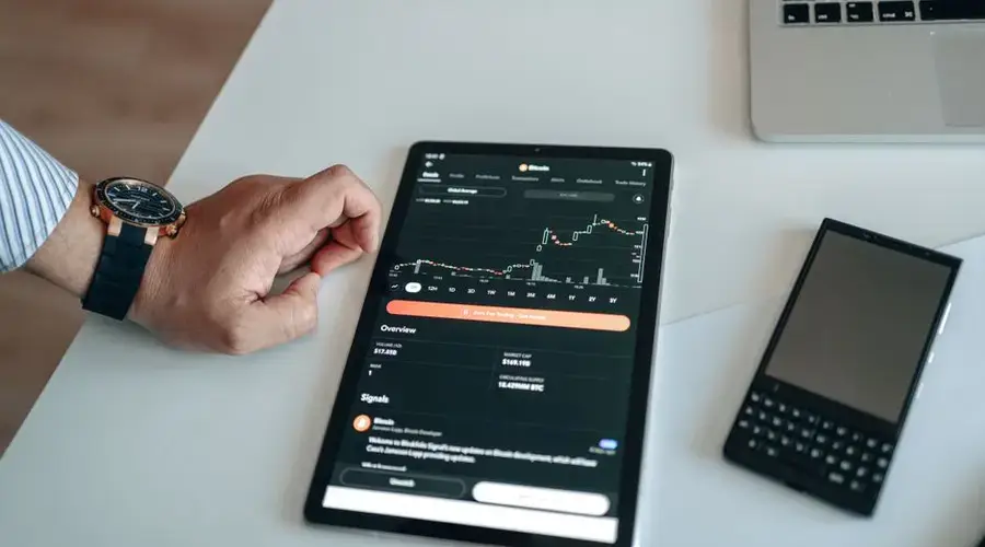 Close-up of a digital tablet displaying a bitcoin chart, with a hand nearby on a white table.
