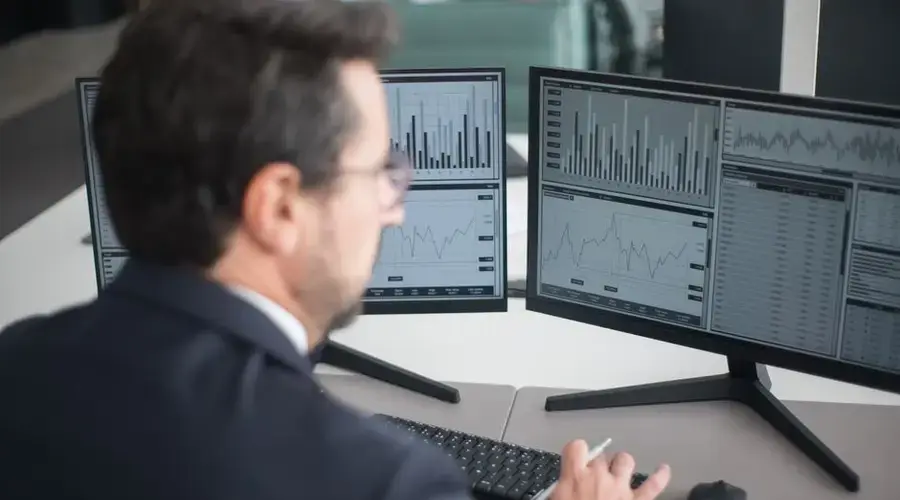 A businessman in a suit analyzing financial charts and graphs on two computer monitors indoors.