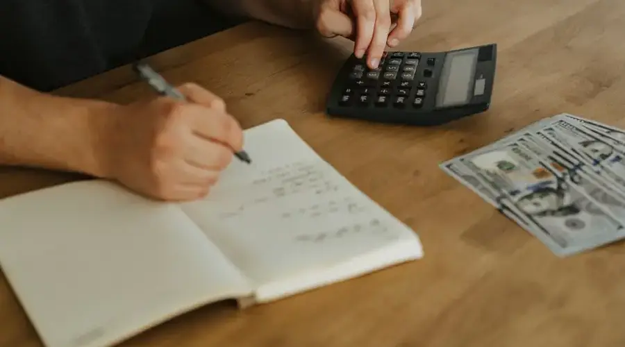 Close-up of hands using a calculator with notebook and cash on a wooden table.
