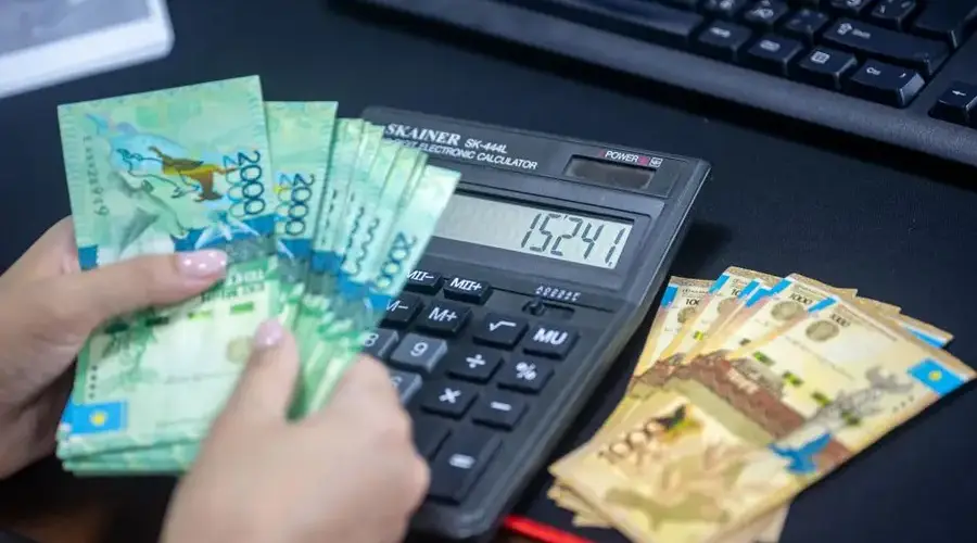 Hands counting Kazakhstani tenge while using calculator at desk.