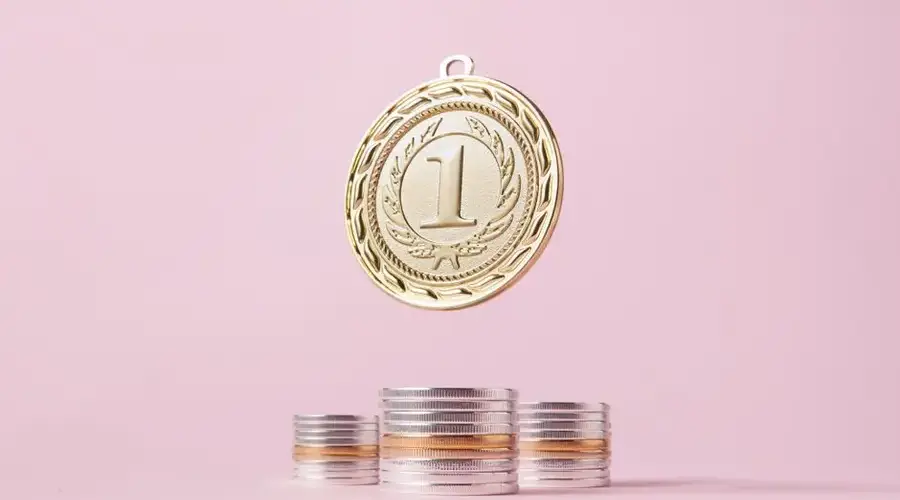 A gold medal and stacks of coins on a light pink backdrop, symbolizing victory and wealth.