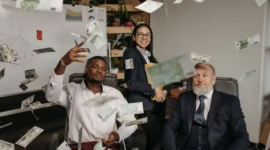 Diverse business team celebrating success with cash raining indoors in office.