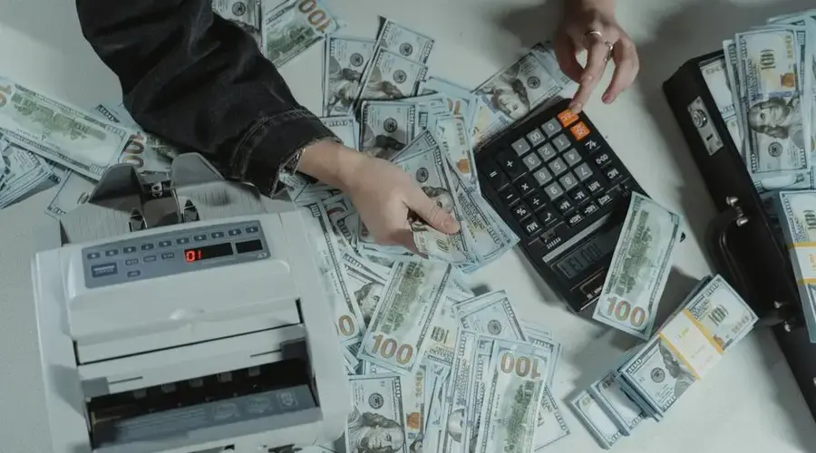 Hands counting 100 dollar bills using a calculator and money counter on a table.
