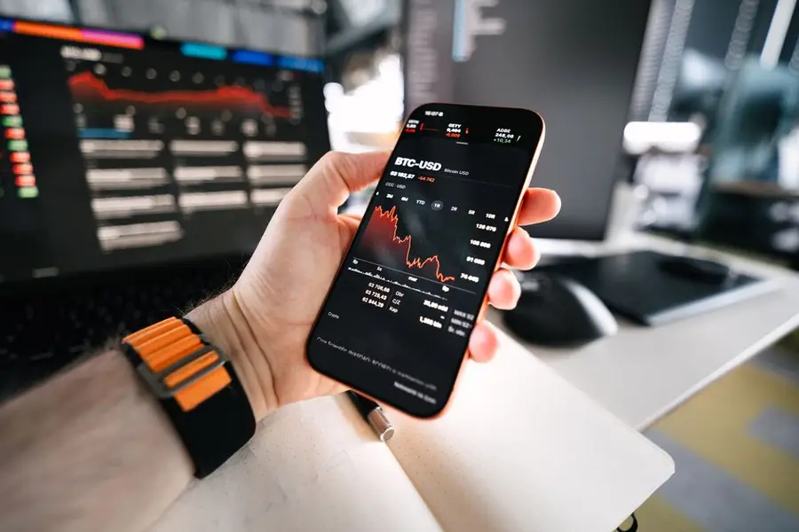 Hand holding smartphone displaying BTC-USD price chart with computer in background.