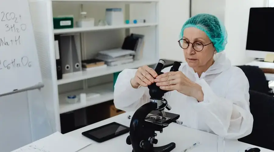 Elderly female scientist in PPE examining samples with a microscope in a modern lab.