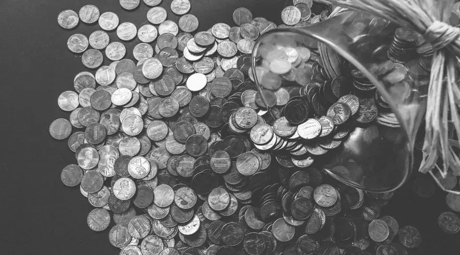 Monochrome image of coins spilling from a glass jar, symbolizing wealth and savings.