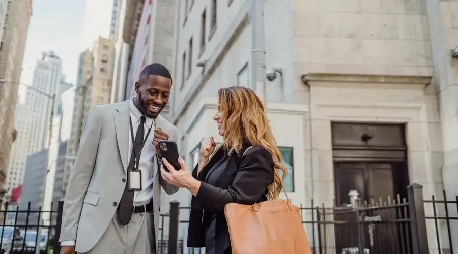 Two business professionals in suits smiling and viewing a cellphone outdoors on Wall Street.