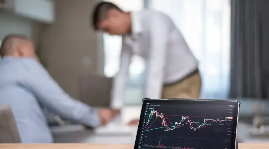 Tablet displaying stock market chart during professional meeting with two men in discussion.