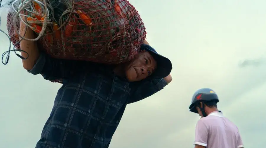 Man carrying a net filled with goods outdoors in daylight, wearing a cap.
