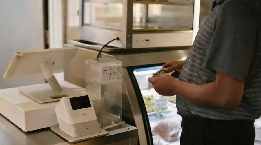 Unrecognizable male in casual clothes standing with money near cash register in grocery store while making purchase near glass showcase