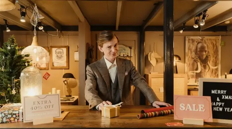 Smiling salesman in a festive store with Christmas decor offering discounts and sales.