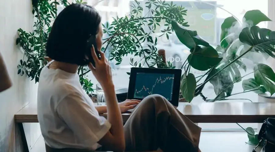 Businesswoman on phone, analyzing stock graphs on laptop in a café.