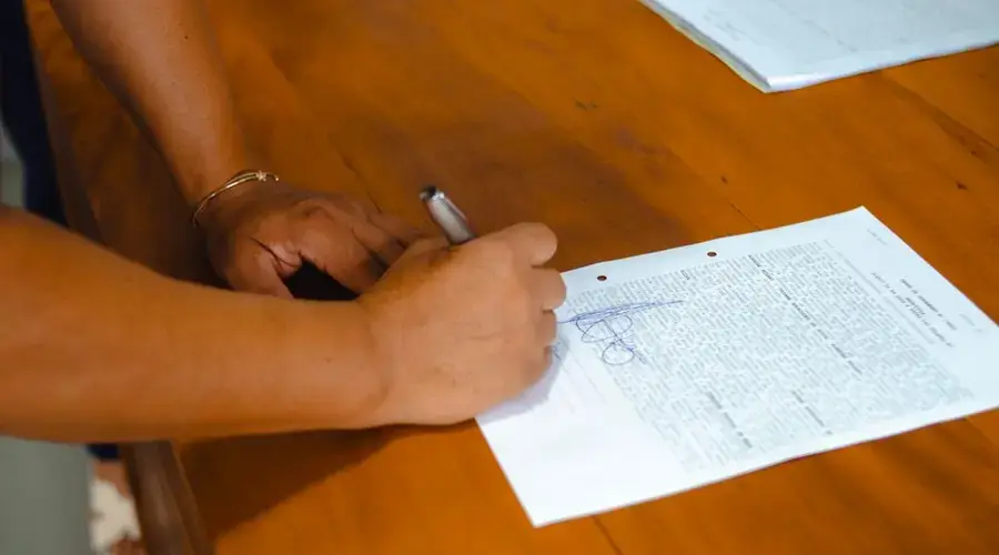 Close-up of hands signing a legal document on a wooden table indoors.
