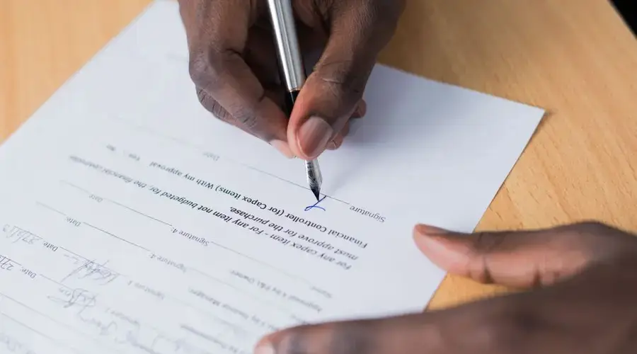A close-up view of a person's hand signing a business contract on a desk with a pen.