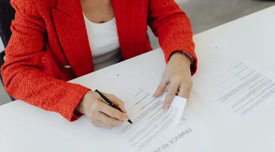 Close-up of a businesswoman in a red blazer signing finance documents at an office desk.