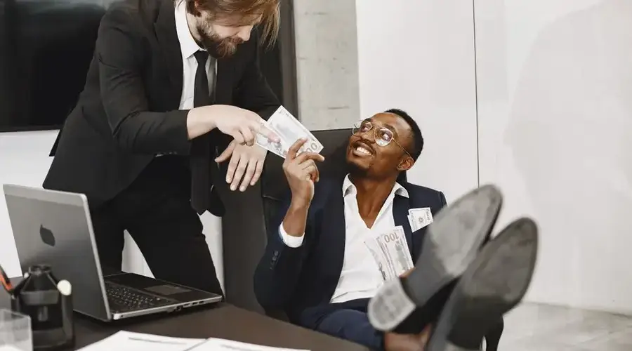 Two businessmen in suits exchanging money with smiles in a modern office environment.