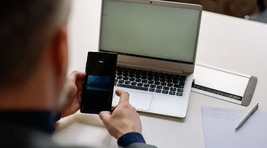 Over the shoulder view of businessman using mobile phone for digital payment beside a laptop in office.