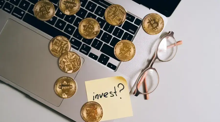Laptop with Bitcoin coins, eyeglasses, and 'invest?' note. Ideal for finance and tech themes.