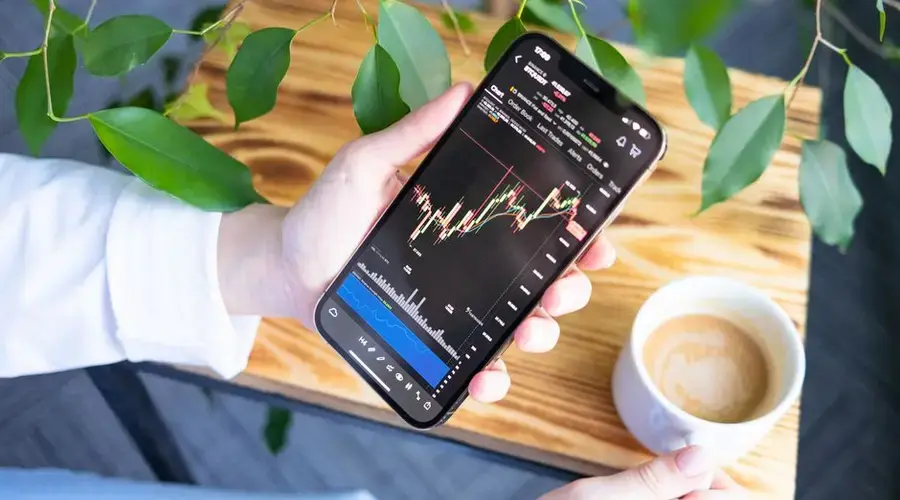 Woman holding smartphone showing stock market graph while enjoying a cup of coffee.