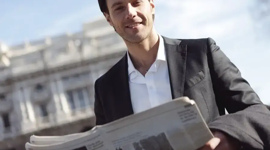 Smiling businessman in suit reads newspaper outside on a sunny day.
