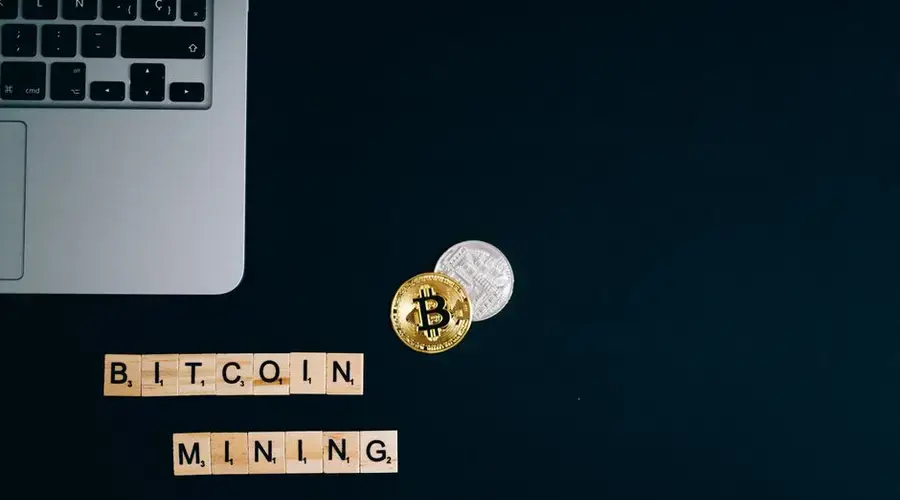 Top view of a laptop with Bitcoin coins and blocks spelling 'Bitcoin Mining' on a dark surface.
