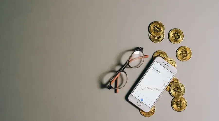 Flat lay of Bitcoin coins, smartphone with graph, and glasses on gray surface.
