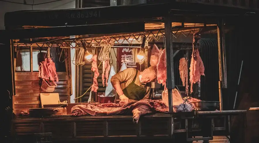 A butcher expertly cuts meat under warm lights at an outdoor night market stall.