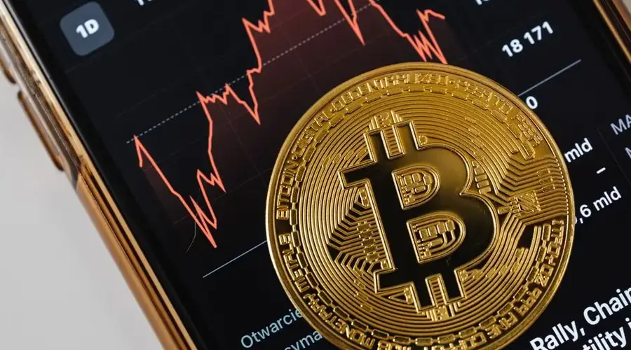 A close-up of a gold Bitcoin coin placed on a smartphone displaying a stock market chart.