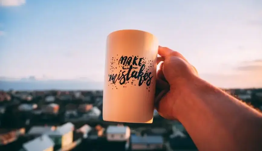 A hand holding a motivational mug with 'Make Mistakes' against a sunset in Lviv.
