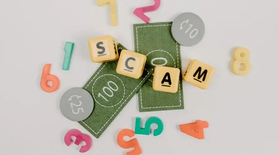 Scrabble tiles spelling 'scam' with play money and numbers on a white background.