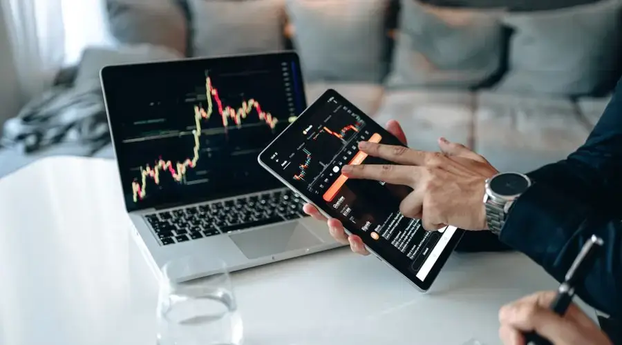 Professional using a tablet for digital trading analysis with a laptop display showing market data.