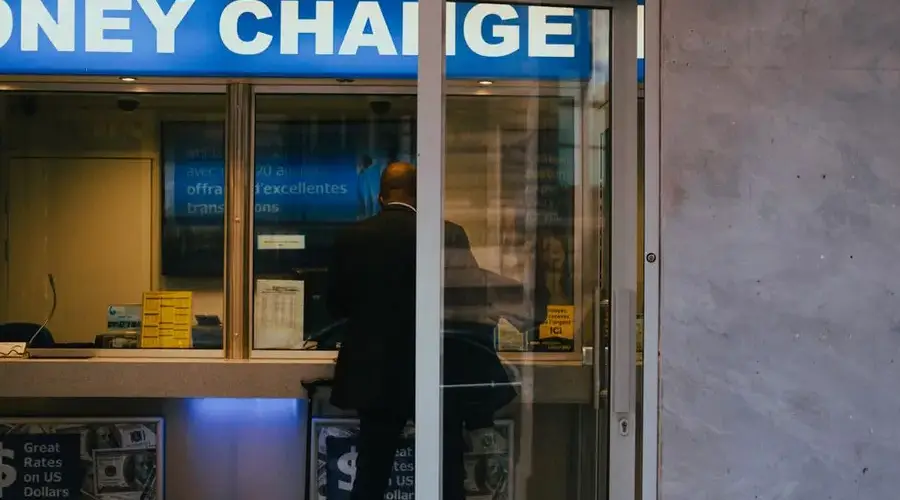 Man at a currency exchange office window, showing currency rates inside a bustling city.