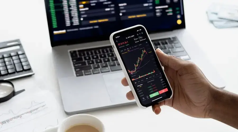 A person trades stocks on a smartphone with a laptop, coffee cup, calculator, and documents in view.
