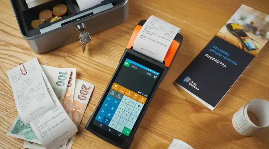 A modern payment terminal with receipts, cash, and manual on a wooden table.