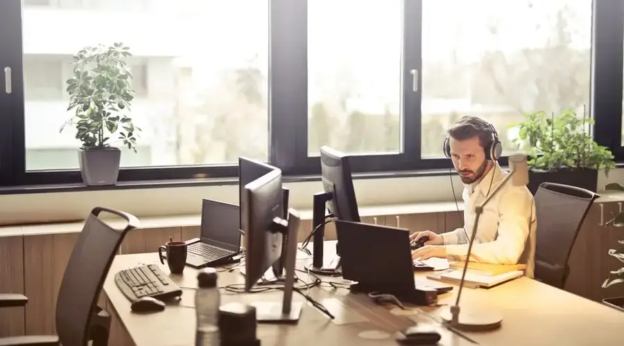 A businessman sits at a desk using multiple computers and a headset in a well-lit modern office.