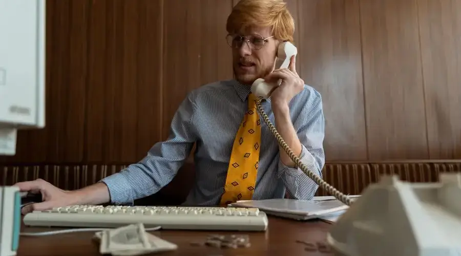 A man in business attire using a vintage telephone and computer in an office setting.