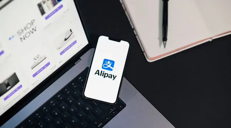 Smartphone displaying Alipay app on open laptop with online shopping site.