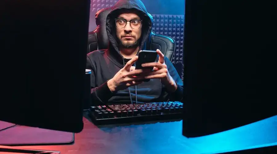 A man in a hoodie using a smartphone, surrounded by tech gear in a dimly lit room.
