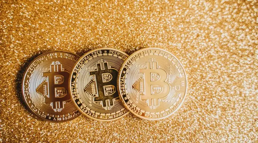 Close-up of three shiny Bitcoin coins on a sparkling gold glitter background, symbolizing cryptocurrency wealth.