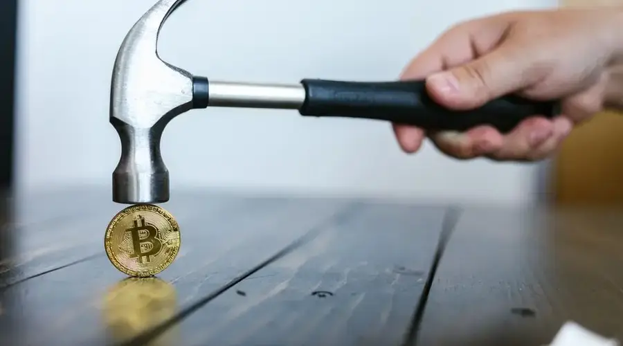 A hand holding a hammer above a Bitcoin coin on a wooden surface, symbolizing cryptocurrency volatility.