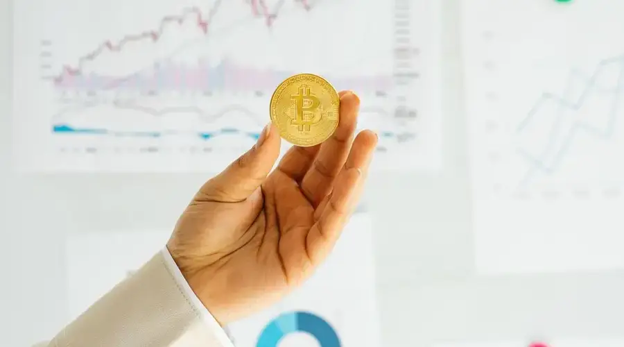 A hand holding a Bitcoin coin against financial charts depicting market trends.
