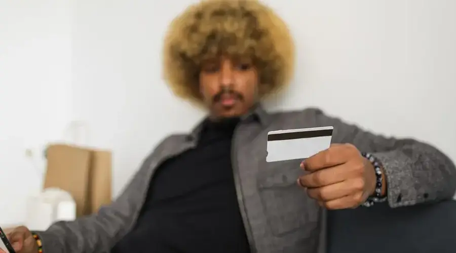 An African American man with afro hair is holding a credit card, making an online purchase indoors.