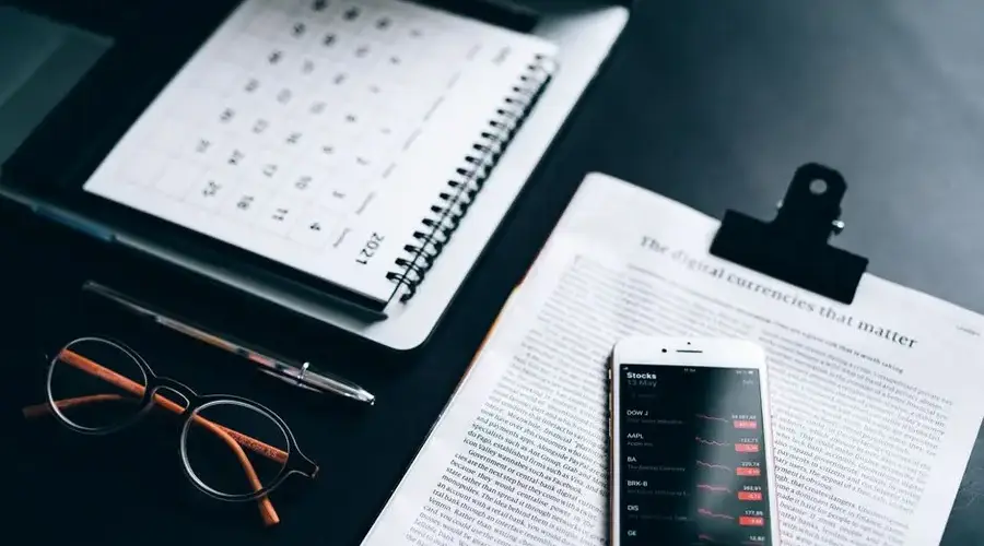 High angle shot of a desk setup with smartphone displaying stock market analytics, clipboard, pen, and glasses.