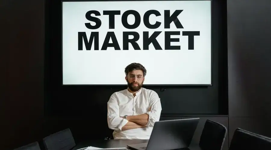 Businessman in modern office setting with a stock market display, looking thoughtful and analytical.
